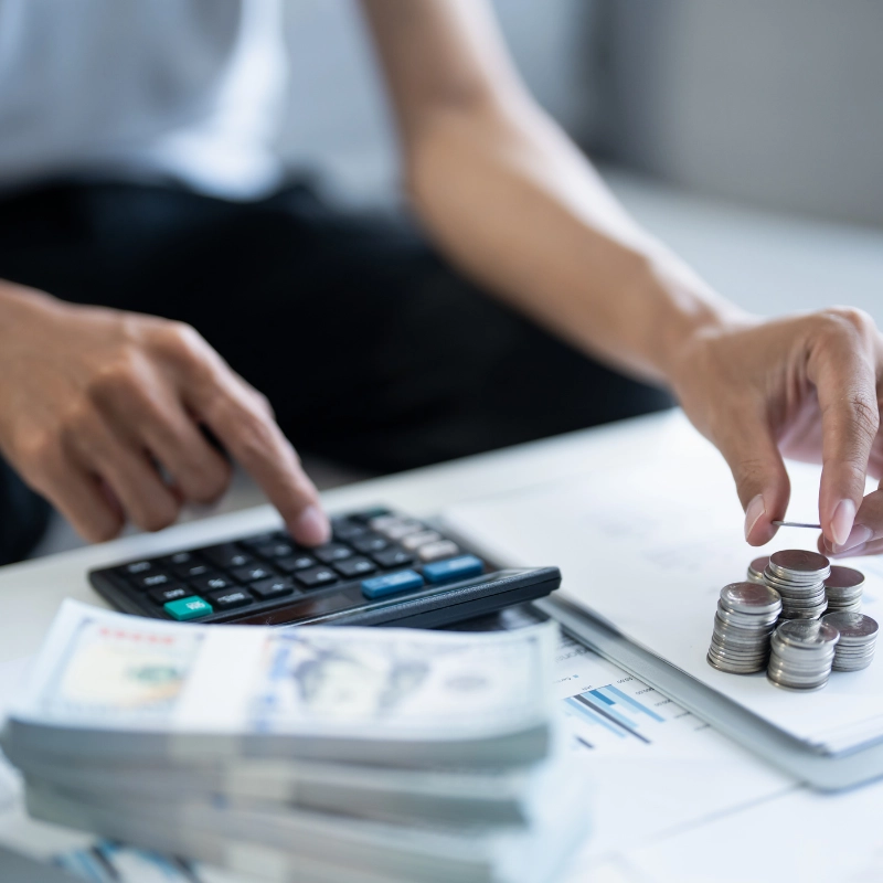 A person uses a calculator with one hand and stacks coins with the other. Bundles of cash and financial documents, including information about the Mass Save Program, are visible on the table, suggesting financial planning or accounting activities.