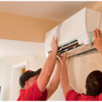 Two workers in red shirts and black caps are performing a mini split installation, servicing a wall-mounted air conditioning unit inside a beige room with natural light streaming through a window.